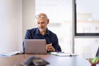 Orta yaşlı işadamı modern bir ofiste oturuyor ve bilgisayarını kullanıyor. Kendine güvenen erkek web konferansı yapıyor..