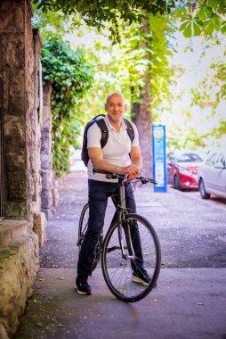 Şehirde bisikletli bir banliyö. Son sınıf öğrencisi sokakta bisiklet sürüyor. Gündelik giysiler ve sırt çantası giyen kendinden emin bir erkek. Tam boy çekim. 