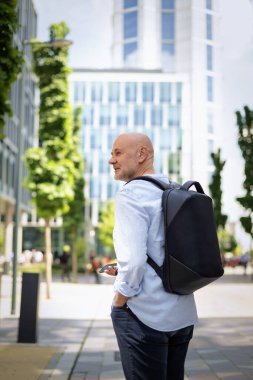Akıllı telefonu olan yakışıklı bir adam ve şehirde finans sektöründe yürüyor. Orta yaşlı iş adamı günlük iş kıyafetleri giyiyor. Arka görünüm. 