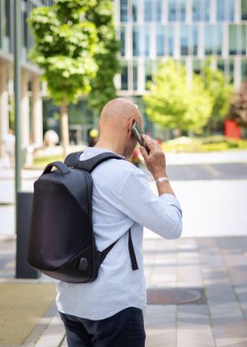 Yakışıklı bir adam dizüstü bilgisayar tutuyor ve şehirde finans sektöründe yürüyor. Orta yaşlı bir işadamı iş kıyafeti giyiyor ve telefonla konuşuyor. Arka görünüm. 