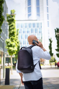 Yakışıklı bir adam dizüstü bilgisayar tutuyor ve şehirde finans sektöründe yürüyor. Orta yaşlı bir işadamı iş kıyafeti giyiyor ve telefonla konuşuyor. Arka görünüm. 