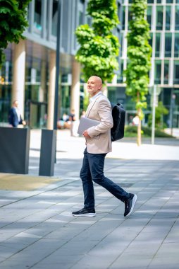 Yakışıklı bir adam dizüstü bilgisayar tutuyor ve şehirde finans sektöründe yürüyor. Orta yaşlı iş adamı günlük iş kıyafetleri giyiyor. Tam boy çekim. 