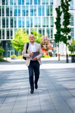 Yakışıklı bir adam dizüstü bilgisayar tutuyor ve şehirde finans sektöründe yürüyor. Orta yaşlı iş adamı günlük iş kıyafetleri giyiyor. Tam boy çekim. 
