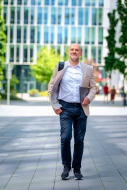 Yakışıklı bir adam dizüstü bilgisayar tutuyor ve şehirde finans sektöründe yürüyor. Orta yaşlı iş adamı günlük iş kıyafetleri giyiyor. Tam boy çekim. 