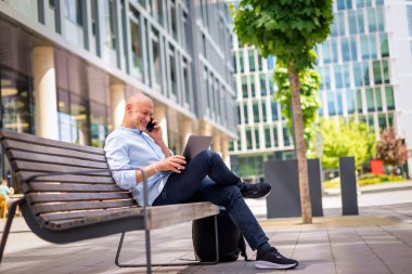 Şehirdeki bir bankta oturan yakışıklı iş adamı cep telefonu ve dizüstü bilgisayar kullanıyor. Profesyonel erkek iş kıyafeti giyiyor ve çalışıyor. Tam boy çekim. 