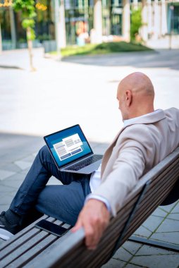 Sokakta bir bankta oturan orta yaşlı bir adamın dizüstü bilgisayarına baktığı dikiz görüntüsü. Ofis dışında çalışan kendine güvenen bir iş adamı..