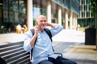 Sokakta bir bankta oturan orta yaşlı bir adam telefon kullanıyor. Gündelik kıyafetler giyen kendinden emin bir erkek.. 