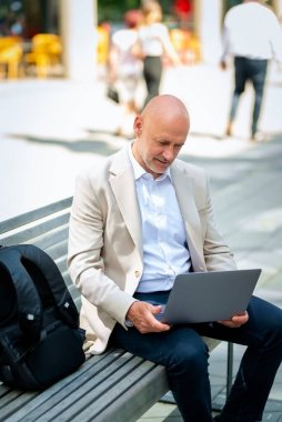 Şehirdeki bir bankta oturup dizüstü bilgisayar kullanan bir iş adamı. Profesyonel erkek iş kıyafeti giyiyor ve çalışıyor..