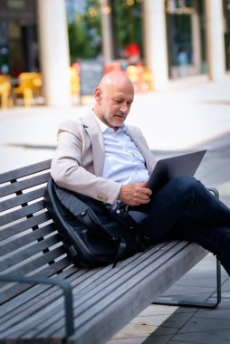 Şehirdeki bir bankta oturup dizüstü bilgisayar kullanan bir iş adamı. Profesyonel erkek iş kıyafeti giyiyor ve çalışıyor..