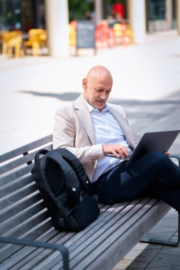 Şehirdeki bir bankta oturup dizüstü bilgisayar kullanan bir iş adamı. Profesyonel erkek iş kıyafeti giyiyor ve çalışıyor..