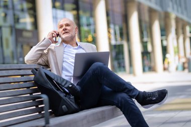 Şehirdeki bir bankta oturan yakışıklı iş adamı cep telefonu ve bilgisayar laptopu kullanıyor. Profesyonel erkek iş kıyafeti giyiyor ve çalışıyor.. 