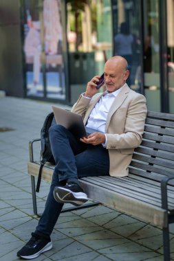 Şehirdeki bir bankta oturan yakışıklı iş adamı cep telefonu ve bilgisayar laptopu kullanıyor. Profesyonel erkek iş kıyafeti giyiyor ve çalışıyor. Tam menzil.. 