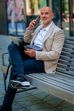 Şehirdeki bir bankta oturan yakışıklı iş adamı cep telefonu ve bilgisayar laptopu kullanıyor. Profesyonel erkek iş kıyafeti giyiyor ve çalışıyor. Tam menzil.. 