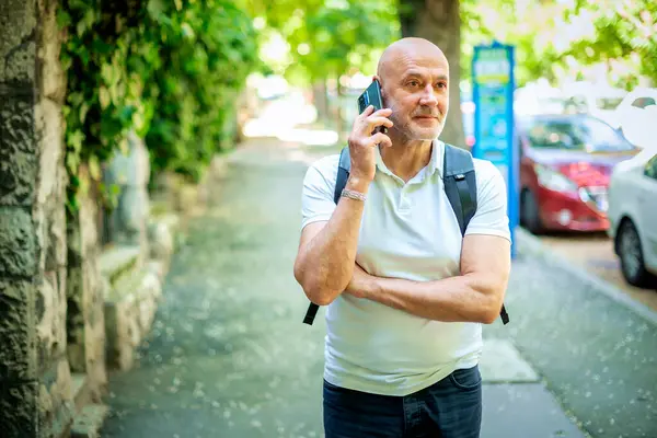Şehirde sokakta yürüyen ve akıllı telefon kullanan orta yaşlı bir adam. Gündelik giysiler ve sırt çantası giyen kendinden emin bir erkek..