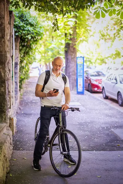 Şehirde bisikletli bir banliyö. Son sınıf öğrencisi sokakta bisiklet sürüyor ve akıllı telefonunu kullanıyor. Gündelik giysiler ve sırt çantası giyen kendinden emin bir erkek. Tam boy çekim. 
