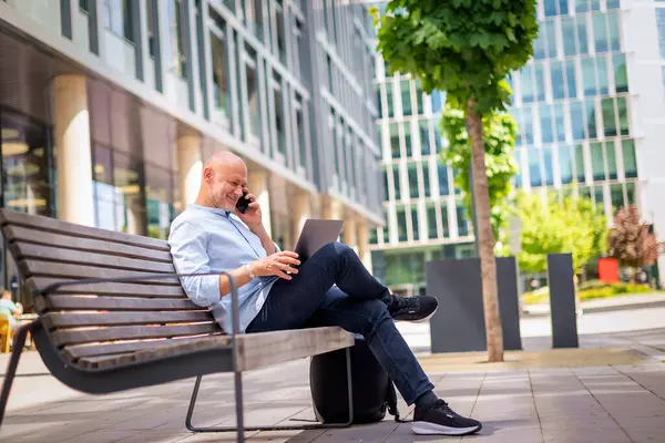 Şehirdeki bir bankta oturan yakışıklı iş adamı cep telefonu ve dizüstü bilgisayar kullanıyor. Profesyonel erkek iş kıyafeti giyiyor ve çalışıyor. Tam boy çekim. 