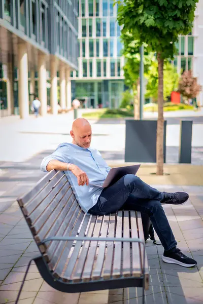 Şehirdeki bir bankta oturup dizüstü bilgisayar kullanan bir iş adamı. Profesyonel erkek, günlük kıyafetler giyiyor ve çalışıyor. Tam boy çekim. 