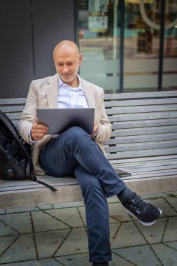 Şehirde bir bankta oturan yakışıklı bir iş adamı ve dizüstü bilgisayar kullanıyor. Profesyonel erkek iş kıyafeti giyiyor ve çalışıyor. Tam boy çekim.