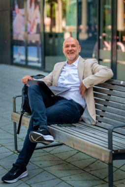 Şehirde bir bankta oturan yakışıklı bir iş adamı ve dizüstü bilgisayar kullanıyor. Profesyonel erkek iş kıyafeti giyiyor ve çalışıyor. Tam boy çekim.