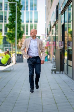 Şehir caddesinde yürüyen orta yaşlı bir adam. Kendine güvenen erkek iş kıyafeti giyiyor. Tam boy çekim. 