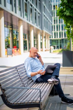Şehirdeki bir bankta oturan yakışıklı iş adamı cep telefonu ve dizüstü bilgisayar kullanıyor. Profesyonel erkek iş kıyafeti giyiyor ve çalışıyor. Tam menzil..