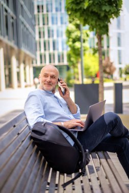 Şehirdeki bir bankta oturan yakışıklı iş adamı cep telefonu ve dizüstü bilgisayar kullanıyor. Profesyonel erkek iş kıyafeti giyiyor ve çalışıyor.. 