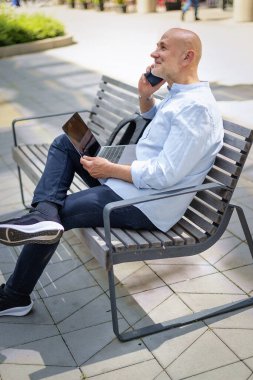 Şehirdeki bir bankta oturan yakışıklı iş adamı cep telefonu ve dizüstü bilgisayar kullanıyor. Profesyonel erkek iş kıyafeti giyiyor ve çalışıyor. Tam menzil..