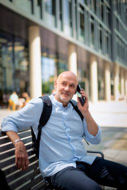 Sokakta bir bankta oturan orta yaşlı bir adam telefon kullanıyor. Gündelik kıyafetler giyen kendinden emin bir erkek.. 