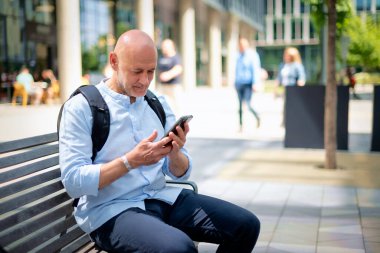 Sokakta bir bankta oturan orta yaşlı bir adam telefon kullanıyor. Gündelik kıyafetler giyen kendinden emin bir erkek.. 