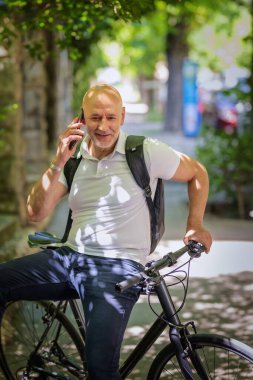 Şehirde bisikletli bir banliyö. Son sınıf öğrencisi sokakta bisiklet sürüyor ve akıllı telefonunu kullanıyor. Gündelik giysiler ve sırt çantası giyen kendinden emin bir erkek. Tam boy çekim. 