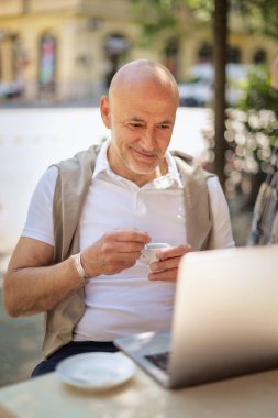 Orta yaşlı bir işadamı bir kafede terasta oturuyor ve laptopuyla çalışıyor. Sıradan kıyafetler giyen profesyonel bir adam.. 
