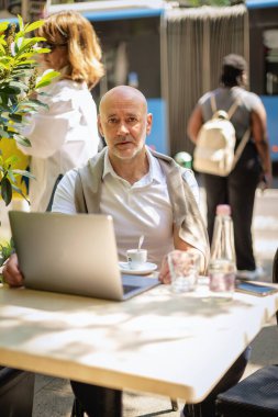 Orta yaşlı bir işadamı bir kafede terasta oturuyor ve laptopuyla çalışıyor. Profesyonel bir adam düşünceli görünüyor.. 