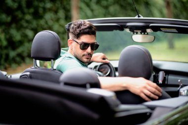 Orta yaşlı bir adam üstü açık bir arabada oturuyor ve araba sürüyor. Gündelik kıyafetler ve güneş gözlüğü takan yakışıklı bir erkek. Orta yaşlı bir adam arabasında oturuyor ve araba sürüyor. Adam arabanın içinde ve geriye bakıyor..