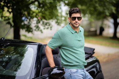Sokakta duran, üstü açık bir arabaya yaslanan orta yaşlı bir adam. Yakışıklı erkek, günlük kıyafetler ve güneş gözlüğü takıyor.. 