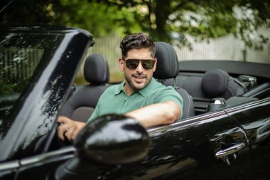 Orta yaşlı bir adam üstü açık bir arabada oturuyor ve araba sürüyor. Yakışıklı erkek, günlük kıyafetler ve güneş gözlüğü takıyor.. 