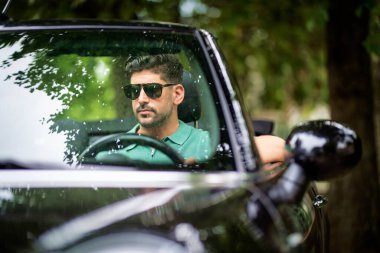 Orta yaşlı bir adam üstü açık bir arabada oturuyor ve araba sürüyor. Yakışıklı erkek, günlük kıyafetler ve güneş gözlüğü takıyor.. 