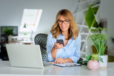 Masada oturan sarı saçlı kadın, bilgisayar ve akıllı telefon kullanıyor. Modern bir ofiste çalışan orta yaşlı bir iş kadını.. 