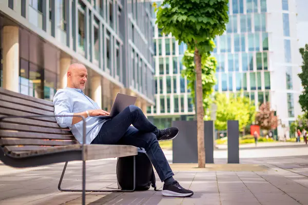 Şehirdeki bir bankta oturan yakışıklı iş adamı cep telefonu ve dizüstü bilgisayar kullanıyor. Profesyonel erkek iş kıyafeti giyiyor ve çalışıyor. Tam menzil..