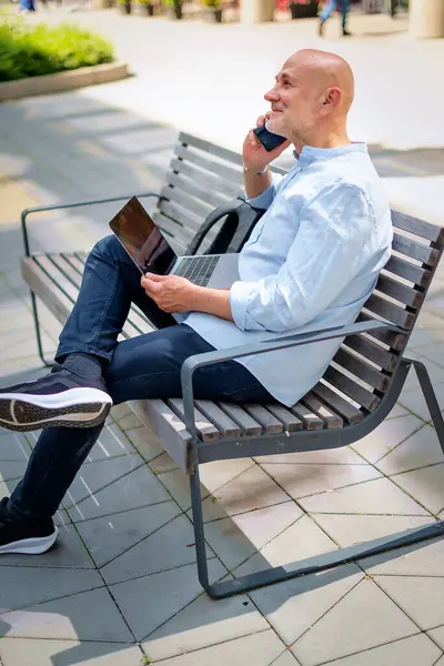 Şehirdeki bir bankta oturan yakışıklı iş adamı cep telefonu ve dizüstü bilgisayar kullanıyor. Profesyonel erkek iş kıyafeti giyiyor ve çalışıyor. Tam menzil..