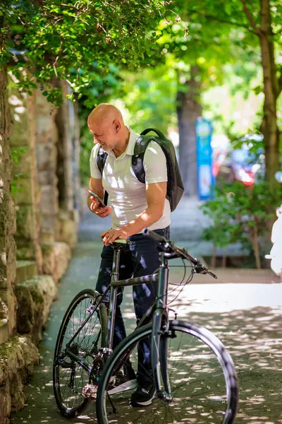 Şehirde bisikletli bir banliyö. Son sınıf öğrencisi sokakta bisiklet sürüyor ve akıllı telefonunu kullanıyor. Gündelik giysiler ve sırt çantası giyen kendinden emin bir erkek. Tam boy çekim.