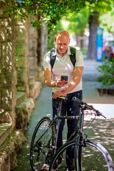 Şehirde bisikletli bir banliyö. Son sınıf öğrencisi sokakta bisiklet sürüyor ve akıllı telefonunu kullanıyor. Gündelik giysiler ve sırt çantası giyen kendinden emin bir erkek. Tam boy çekim.