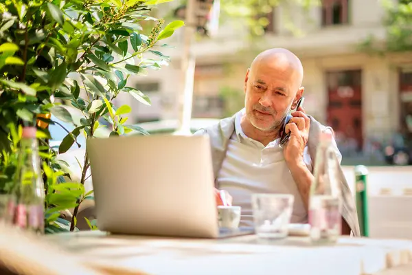Orta yaşlı bir işadamı bir kafede terasta oturuyor ve laptopuyla çalışıyor. Gündelik kıyafetler giyen ve telefonla konuşan profesyonel bir adam..
