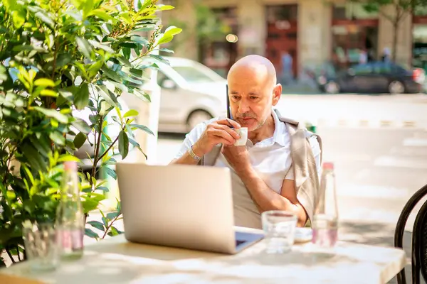Orta yaşlı bir işadamı bir kafede terasta oturuyor ve laptopuyla çalışıyor. Gündelik kıyafetler giyen ve telefonla konuşan profesyonel bir adam..