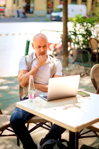 Orta yaşlı bir işadamı bir kafede terasta oturuyor ve laptopuyla çalışıyor. Profesyonel bir adam perdeye özenle bakıyor. Eller çenede..  