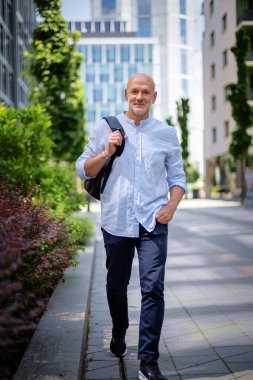 Şehir caddesinde yürüyen orta yaşlı bir adam. Tişört ve sırt çantası giyen kendinden emin bir erkek. Tam boy çekim.