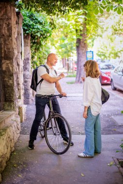Bisikletinin yanında duran ve sokakta bir kadınla konuşan bir adam. Orta yaşlı bir adam ve günlük kıyafetler giyen sarışın bir kadın. Tam uzunluk. 