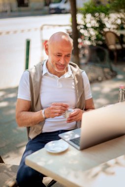 Orta yaşlı bir işadamı bir kafede terasta oturuyor ve laptopuyla çalışıyor. Gündelik kıyafetler giyen ve kahve içen profesyonel bir adam..  
