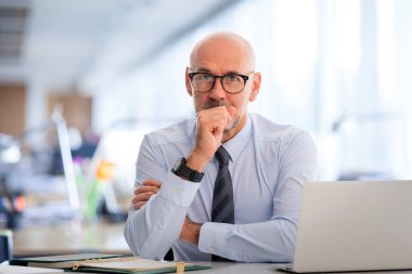 Ofisteki masasında dizüstü bilgisayarıyla çalışan orta yaşlı bir iş adamı. Profesyonel erkek, düşünceli bir şekilde kameraya bakıyor..