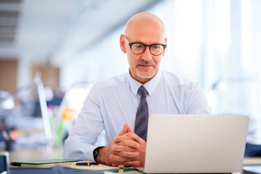 Ofisteki masasında dizüstü bilgisayarıyla çalışan orta yaşlı bir iş adamı. Profesyonel erkek, özenle dizüstü bilgisayara bakıyor..