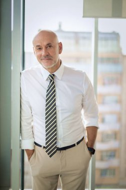 Ofiste dikilip kameraya bakan orta yaşlı bir iş adamı. Gömlek ve kravat takan kendinden emin profesyonel bir adam.. 